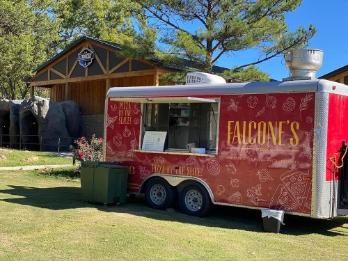A red food truck named "Falcone's" serving pizza, parked near a wooden building and surrounded by greenery.