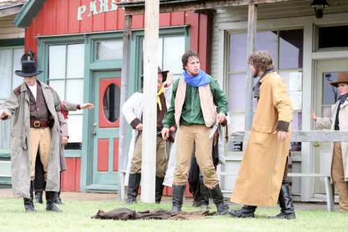 A group of people in Western attire stands outside a building, appearing to engage in a dramatic scene.