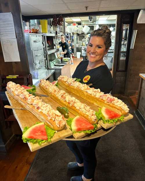 A smiling woman holds a tray of large lobster rolls with lettuce and watermelon slices in a restaurant kitchen.