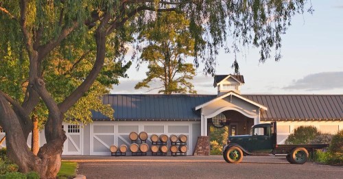 A vintage truck parked in front of a winery with barrels and a charming building under a clear sky.