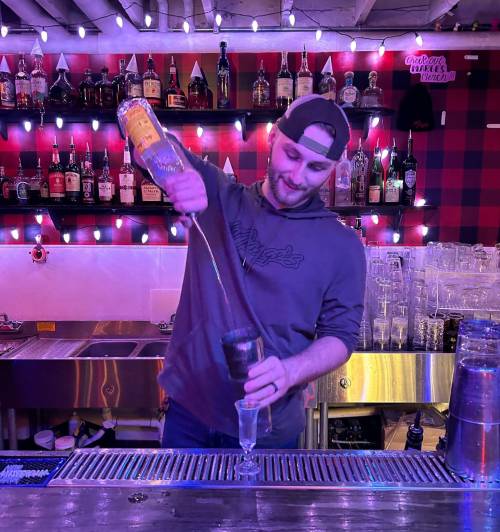 A bartender pours a drink into a glass at a vibrant bar with colorful lights and shelves of liquor in the background.
