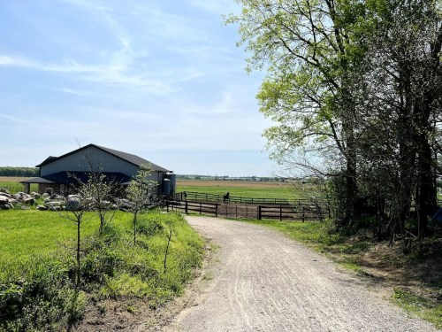 A gravel path leads to a blue barn, surrounded by green fields and trees under a clear blue sky.