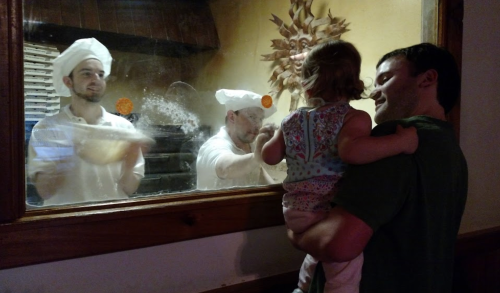 A man holds a child while watching two chefs making pizza through a window.