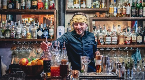 A bartender in a black shirt smiles while mixing drinks at a well-stocked bar with various bottles and ingredients.