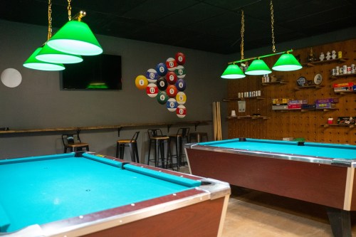A pool hall featuring two tables, green pendant lights, and a wall decorated with colorful billiard balls.