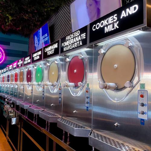A row of colorful slushy machines with various flavors, including cookies and milk, displayed in a vibrant setting.