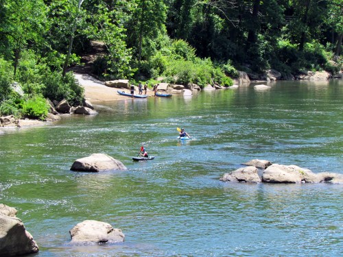 Two people kayaking on a river, with a sandy shore and trees in the background. Others are gathered on the bank.