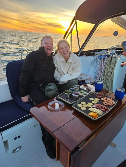 A couple smiles on a boat at sunset, enjoying a platter of snacks and drinks. The ocean glistens in the background.