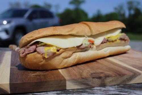 A large sub sandwich with roast beef, cheese, and toppings, resting on a wooden cutting board outdoors.