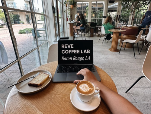 A laptop displaying "Reve Coffee Lab" next to a slice of cake and a latte on a wooden table in a cozy café.