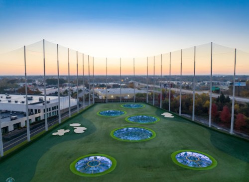 Aerial view of a golf driving range with circular targets, surrounded by netting and a sunset in the background.