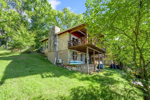 A two-story stone house surrounded by greenery, featuring a balcony and outdoor seating on a sunny day.