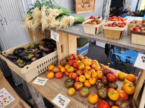 A market stall displays fresh vegetables, including eggplants, tomatoes, and green onions, in rustic wooden crates.