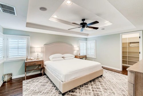 Bright, modern bedroom with a queen bed, ceiling fan, and light-colored walls, featuring a cozy rug and plantation shutters.