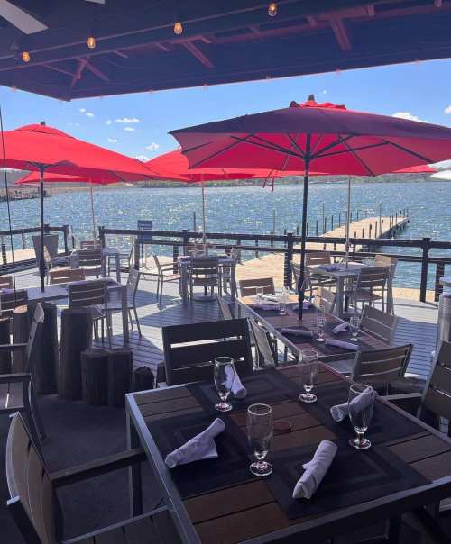 Outdoor dining area with red umbrellas overlooking a lake, featuring tables set for guests and a wooden dock in the background.