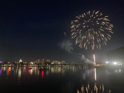 Colorful fireworks illuminate the night sky over a city skyline, reflecting on a calm lake.