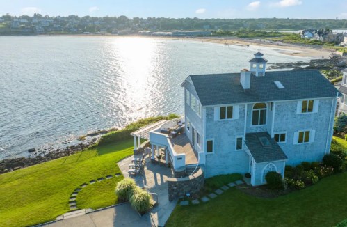 Aerial view of a coastal home with a patio, overlooking a shimmering bay and sandy beach in the background.