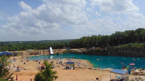 A scenic view of a blue swimming area surrounded by sandy beaches and green trees under a partly cloudy sky.