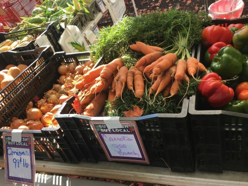 Baskets of fresh vegetables at a market, including carrots, onions, and bell peppers, with price signs.