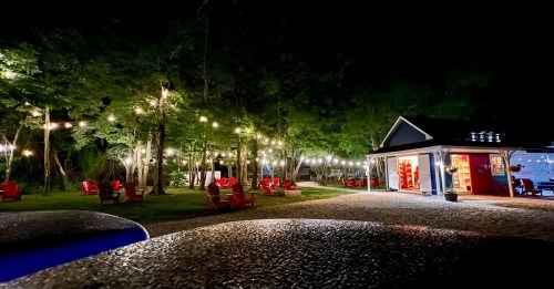 A cozy outdoor setting at night with string lights, red chairs, and a brightly lit building surrounded by trees.