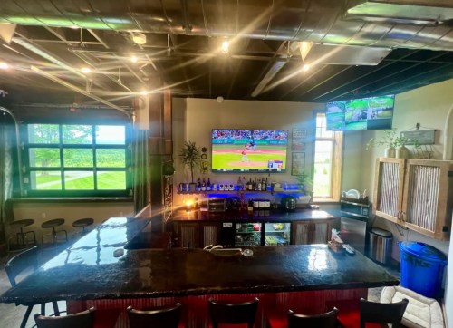 A modern bar with a large screen showing a baseball game, surrounded by stools and a view of greenery outside.
