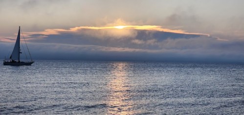 A sailboat glides across calm waters as the sun sets behind clouds, casting a warm glow on the horizon.