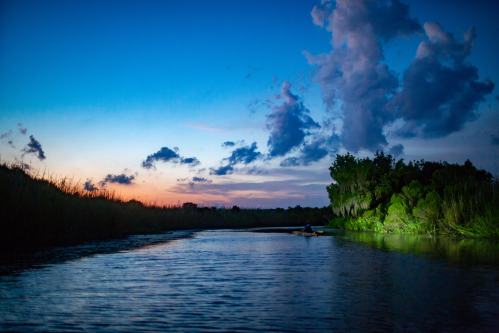 A serene river scene at dusk, with vibrant clouds and lush greenery reflecting in the calm water.