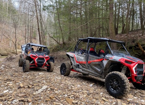 Two red and black off-road vehicles parked on a rocky path in a wooded area. Trees line the background.