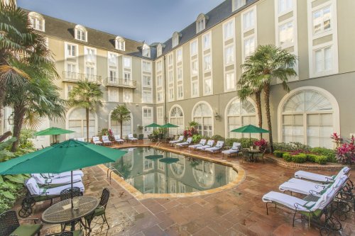 A serene hotel courtyard featuring a pool surrounded by lounge chairs and tropical plants under green umbrellas.