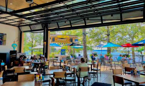 A lively restaurant with indoor and outdoor seating, featuring umbrellas and a view of water and trees.