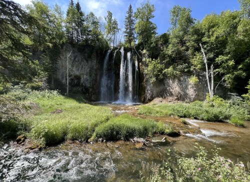 A serene waterfall cascades down rocky cliffs, surrounded by lush greenery and a clear stream in a sunny landscape.