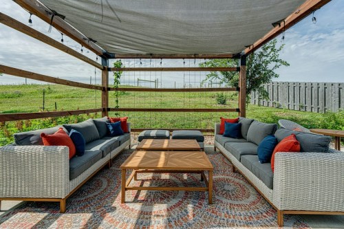Cozy outdoor seating area with gray sofas, colorful pillows, and a wooden table, surrounded by greenery and a cloudy sky.