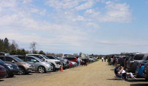 A gravel parking lot filled with cars, with people sitting and walking under a partly cloudy sky.