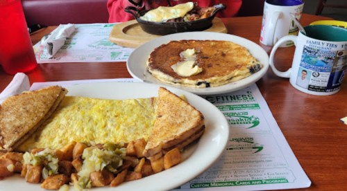 A breakfast spread featuring an omelet, toast, home fries, and a pancake with butter, alongside a coffee mug.