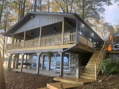 A modern lakeside house with a spacious deck, surrounded by trees and featuring stairs leading to the entrance.