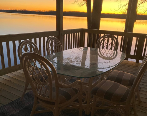 A glass table with six chairs on a wooden deck overlooking a calm lake at sunset.