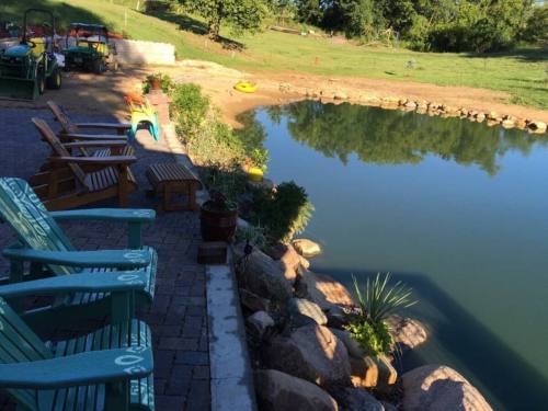 A serene pond view with wooden chairs, rocks, and greenery, surrounded by a grassy area and kayaks on the water.