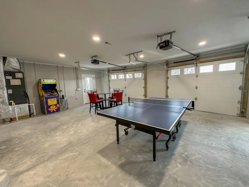 A spacious garage with a ping pong table, red chairs, and an arcade game in the corner. Bright and well-lit.