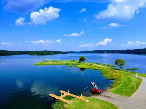 A serene lake view with a wooden dock, a red car, and lush greenery under a blue sky with fluffy clouds.
