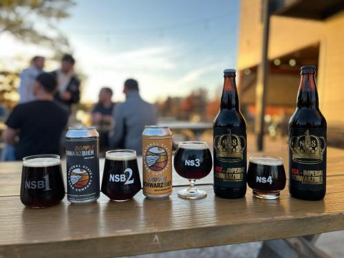 A variety of craft beers in cans and bottles on a wooden table, with people socializing in the background.