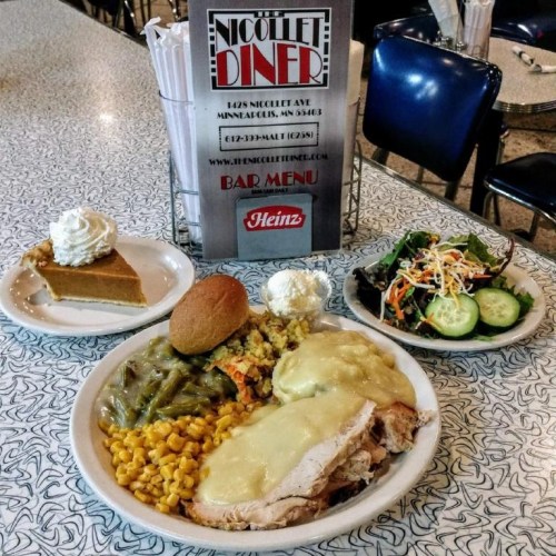 A plate of turkey with gravy, corn, green beans, stuffing, a roll, and a side salad, with a dessert and diner menu in the background.