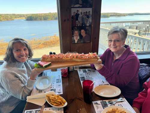 Two women smile while holding a large seafood sub on a wooden platter, with a scenic view of a lake in the background.