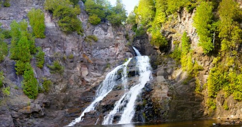 A scenic waterfall cascading down rocky cliffs, surrounded by lush green trees and a calm pool at the base.
