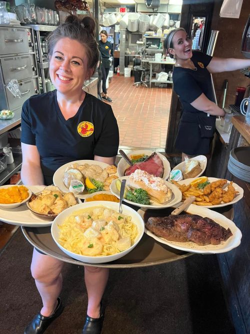 A waitress carries a large tray of various dishes in a busy restaurant kitchen. Another staff member smiles in the background.