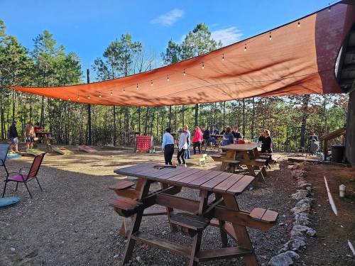 Outdoor gathering space with picnic tables, string lights, and a shaded area under a large canopy in a wooded setting.