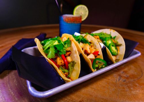 Three tacos filled with grilled chicken, peppers, and cilantro, served on a metal tray with a colorful drink in the background.