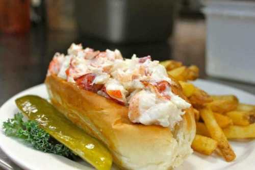 A lobster roll topped with fresh lobster meat, served with fries and a pickle on a white plate.