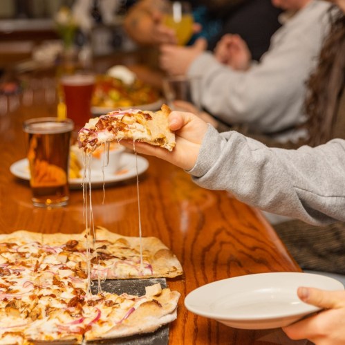 A hand reaches for a cheesy pizza slice at a lively table with drinks and people enjoying a meal together.