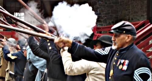 A group of soldiers in historical uniforms firing muskets, with smoke billowing from the barrels.