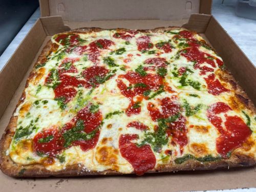 A rectangular pizza with melted cheese, red sauce, and green pesto, served in a cardboard box.
